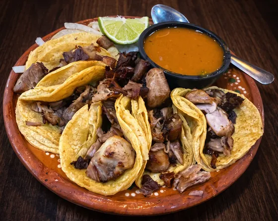 Un plato de barro con tacos de carnitas, trozos de carne, cebolla, un gajo de lim&oacute;n y un taz&oacute;n de consom&eacute; con su cuchara, sobre una mesa de madera oscura.