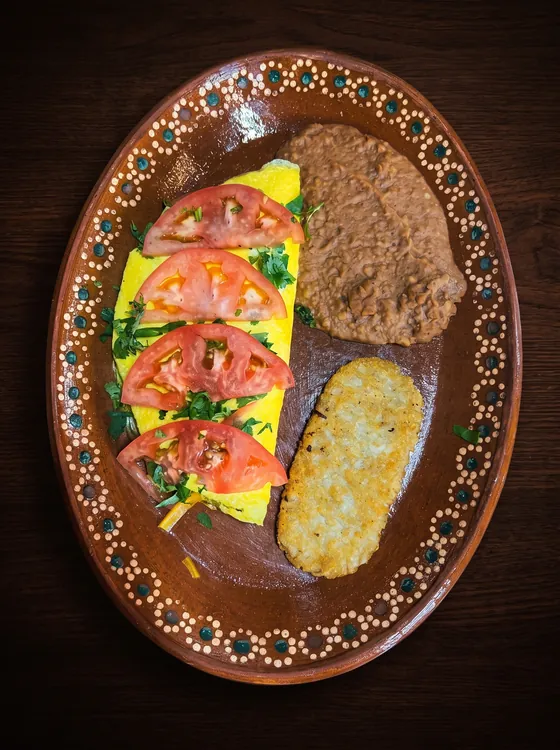 A yellow omelette topped with tomato slices and cilantro, beside refried beans and a golden hash brown on a clay plate.