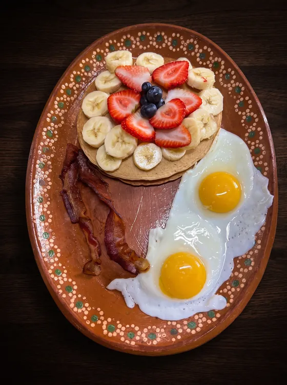 A stack of pancakes topped with strawberries, banana slices and blueberries, with two fried eggs and strips of bacon.