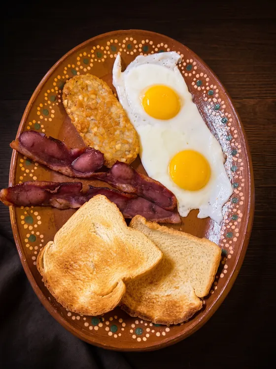 Two fried eggs with strips of bacon, a hash brown and two slices of toast on a clay plate.