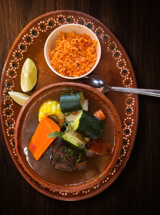 A clay bowl of beef soup with corn, courgette and carrot, with a cup of rice, lime wedges and a spoon on a clay plate.