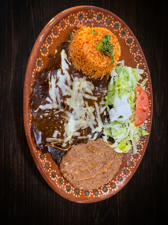 Enchiladas under dark mole and melted cheese beside rice, refried beans, lettuce, sour cream and tomato.