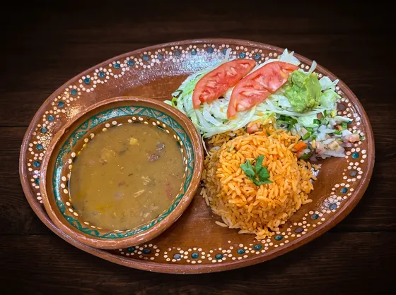 A clay bowl of soup beside rice with a sprig of cilantro, lettuce, guacamole and tomato.
