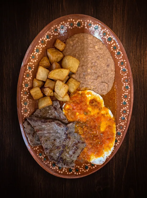 Thin grilled steak and eggs under red sauce, with fried potato cubes and refried beans.