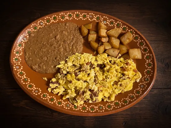 Scrambled eggs beside fried potato cubes and refried beans on an oval clay plate.