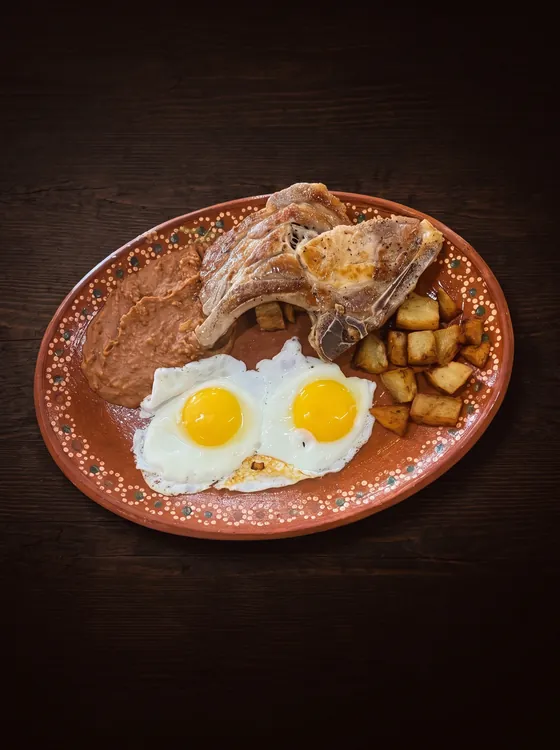 A grilled bone-in chop with two fried eggs, fried potato cubes and refried beans.
