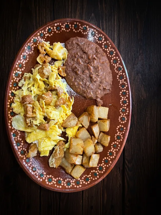Scrambled eggs with pieces of fried pork rind, beside fried potato cubes and refried beans.