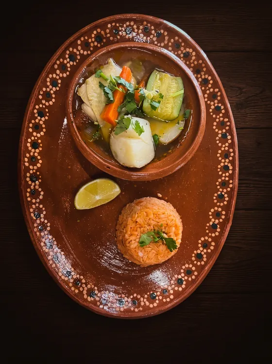 A clay bowl of vegetable soup with potato, courgette and carrot, beside a mound of rice and a lime wedge.
