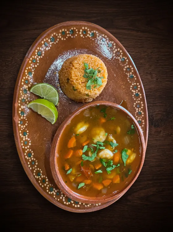A clay bowl of orange broth with vegetables and cilantro, beside a mound of rice and lime wedges.