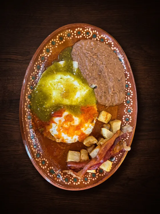 Two fried eggs, one under green salsa and one under red, with fried potatoes, bacon and refried beans.