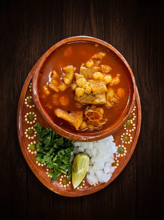 A clay bowl of red broth with tripe and hominy, served with chopped onion, cilantro and a lime wedge.