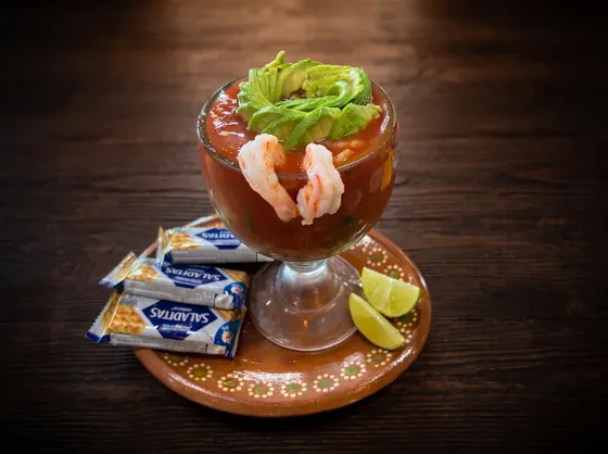 Camarones y aguacate en una copa con salsa roja, sobre un plato de barro con limones y tres paquetes de galletas saladas.