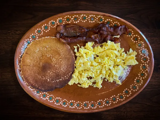 A thick golden round of fried masa beside scrambled eggs and strips of bacon.