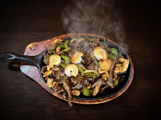 A sizzling cast-iron skillet on a wooden board, steaming with grilled beef, chicken, shrimp, peppers and onion.