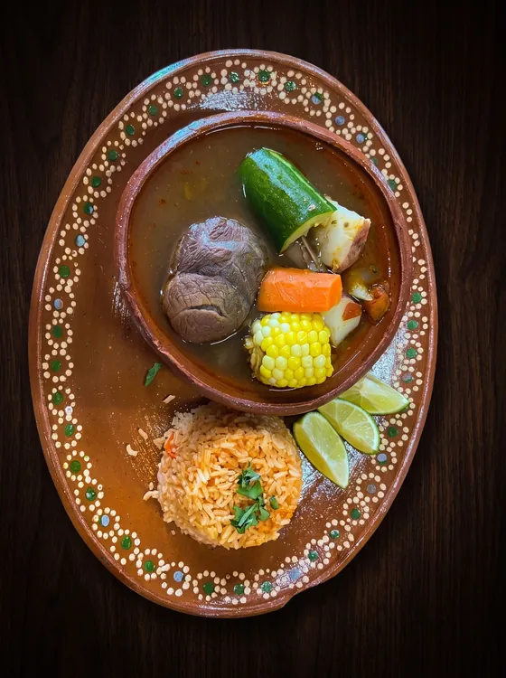 A clay bowl of beef soup with corn, carrot, courgette and potato, beside rice and lime wedges.