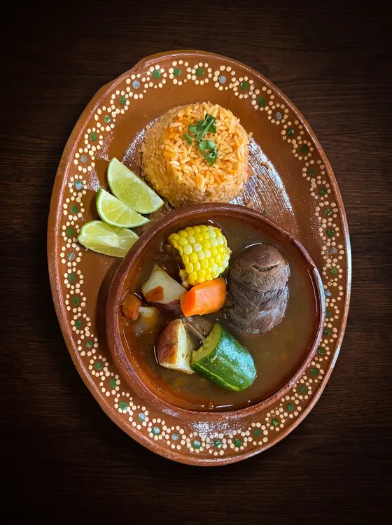 Beef soup with corn on the cob, courgette and carrot in a clay bowl, beside rice and lime wedges.