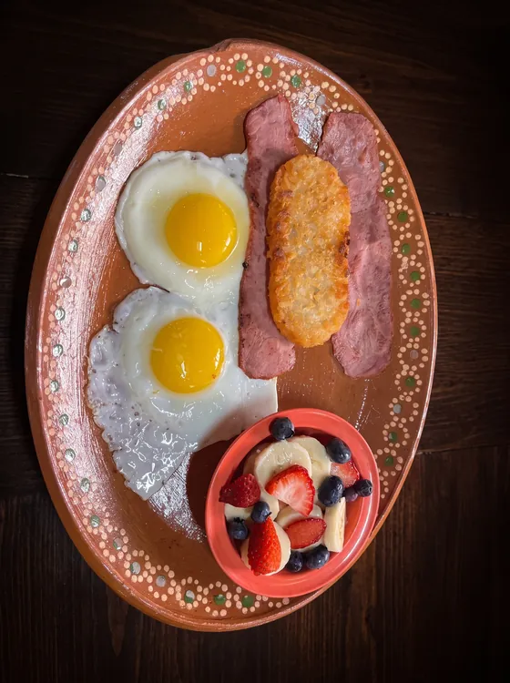 Two fried eggs with slices of ham, a hash brown and a cup of strawberries, banana and blueberries.
