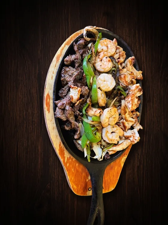 Sartén de hierro sobre una tabla de madera con tiras de res, camarones, pollo y chile verde.