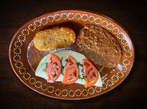 A pale folded dish topped with tomato slices and cilantro, beside refried beans and a golden hash brown.