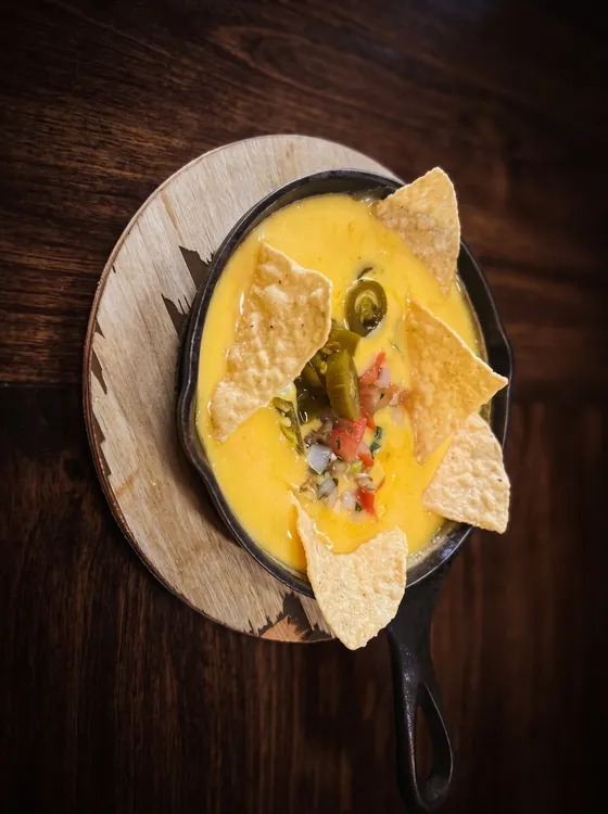 Queso amarillo derretido en un sartén de hierro con totopos, jalapeño y pico de gallo, sobre una tabla de madera.