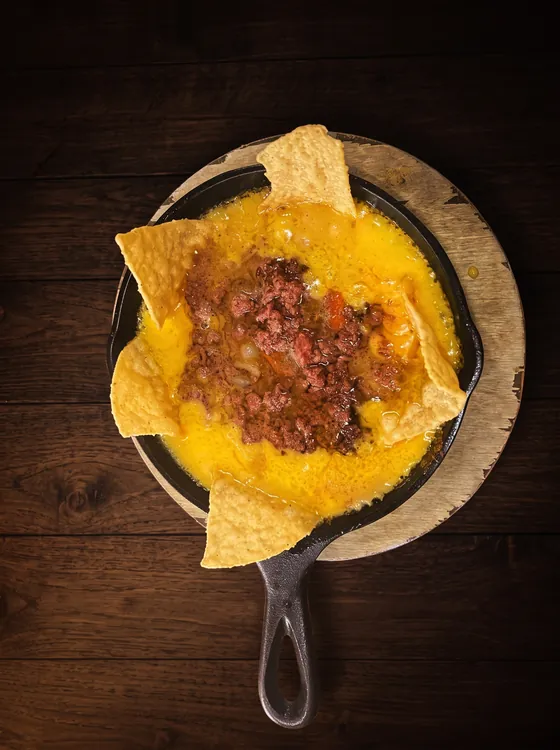 Melted yellow cheese with seasoned minced beef in a cast-iron skillet, ringed with tortilla chips, on a wooden board.