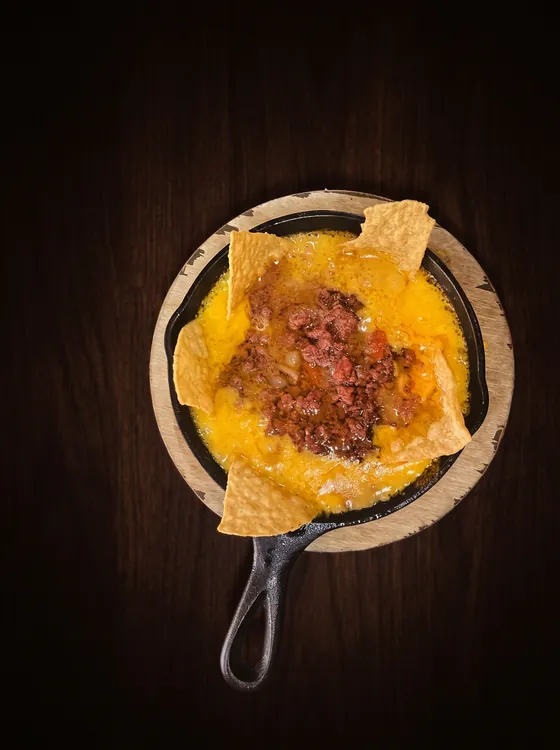 A second view: melted cheese with seasoned minced beef in a cast-iron skillet, with tortilla chips, on a wooden board.