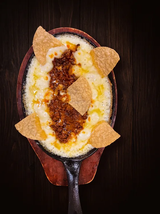 Melted white cheese with chorizo in a cast-iron skillet, topped with tortilla chips, on a wooden board.