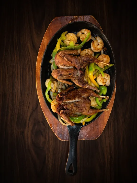 A cast-iron skillet on a wooden board with two whole grilled quail, shrimp, peppers and onion.