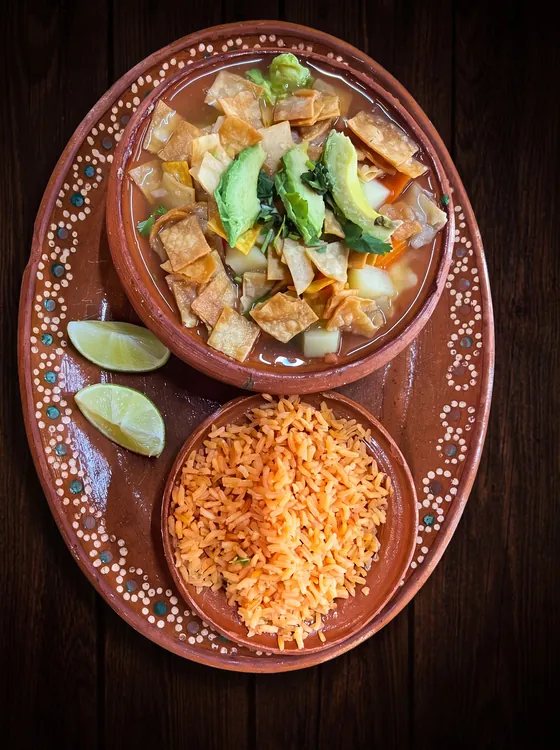 A clay bowl of broth with fried tortilla strips and avocado slices, beside a bowl of rice and lime wedges.