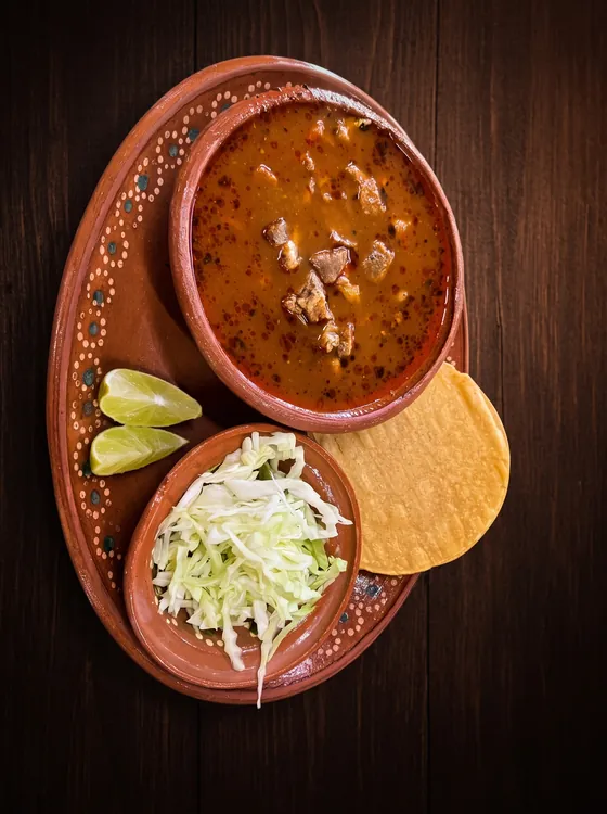 A clay bowl of red broth with pieces of meat, served with shredded cabbage, a crisp tostada and lime wedges.