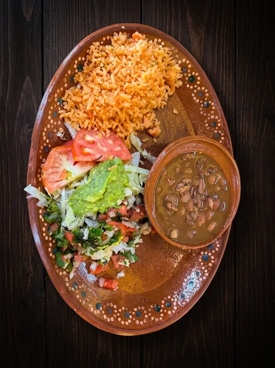 Rice, a cup of whole beans in broth, guacamole, pico de gallo, lettuce and tomato on a clay plate.