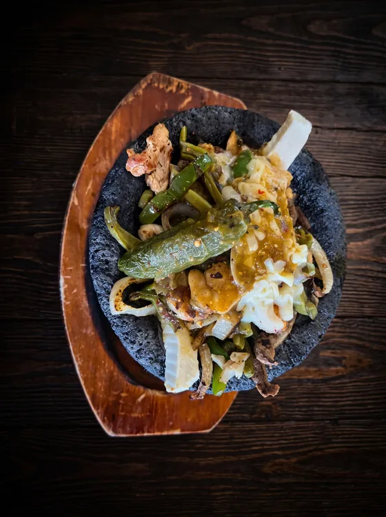 A lava-stone bowl on a wooden board filled with grilled meats, melted cheese, cactus and a roasted jalapeño.