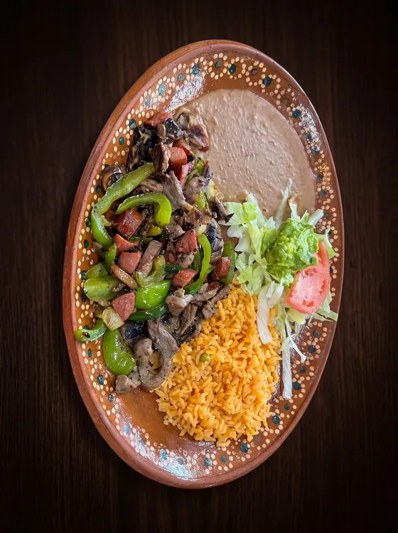 Grilled beef strips with sausage, green pepper and onion, beside refried beans, rice, lettuce, guacamole and tomato.